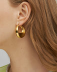 Close-up of a woman's ear adorned with a chunky oval design, 18kt gold-plated **Moon Earrings Gold by For Art's Sake®**. The shiny earring reflects light, highlighting its polished surface. The woman has light brown hair, and visible strands frame the ear. She is wearing a light green top.
