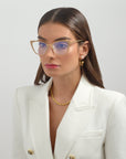 A person with long brown hair is wearing For Art's Sake® Stella Two gold-rimmed optic glasses, a white blazer, and gold jewelry, including hoop earrings and a necklace. They look to the side against a plain background.
