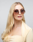 A woman with long, blonde hair is wearing large, stylish For Art's Sake® Palace sunglasses with 18-karat gold-plated frames and brown-tinted, UVA & UVB-protected lenses. She is dressed in a light beige outfit and is looking slightly to the side with a neutral expression. The background is plain and light gray.
