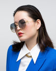A person with straight, dark hair is wearing large, UVA & UVB-protected For Art's Sake® Palace gold-rimmed sunglasses with gradient lenses. They are dressed in a blue blazer over a white collared shirt and accessorized with 18-karat gold-plated chain earrings and necklace. The background is a solid light gray.