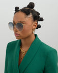A stylish woman with Bantu knots wears For Art's Sake® Birthday Cake-Blue oversized sunglasses, a green blazer, and unique gold and faux pearl earrings against a plain light background.