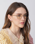 The Lady-S by For Art's Sake® complements the woman with long brown hair. Her chic cat-eye glasses enhance her timeless elegance as she gazes sideways in a yellow crocheted sweater over a beige bow blouse against a plain background.