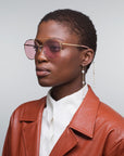 A person wearing For Art's Sake® Yoyo - ARC pink-tinted aviator sunglasses with 18-karat gold plating, long detachable pearl earrings, and a brown leather jacket over a white shirt, set against a neutral gray background.