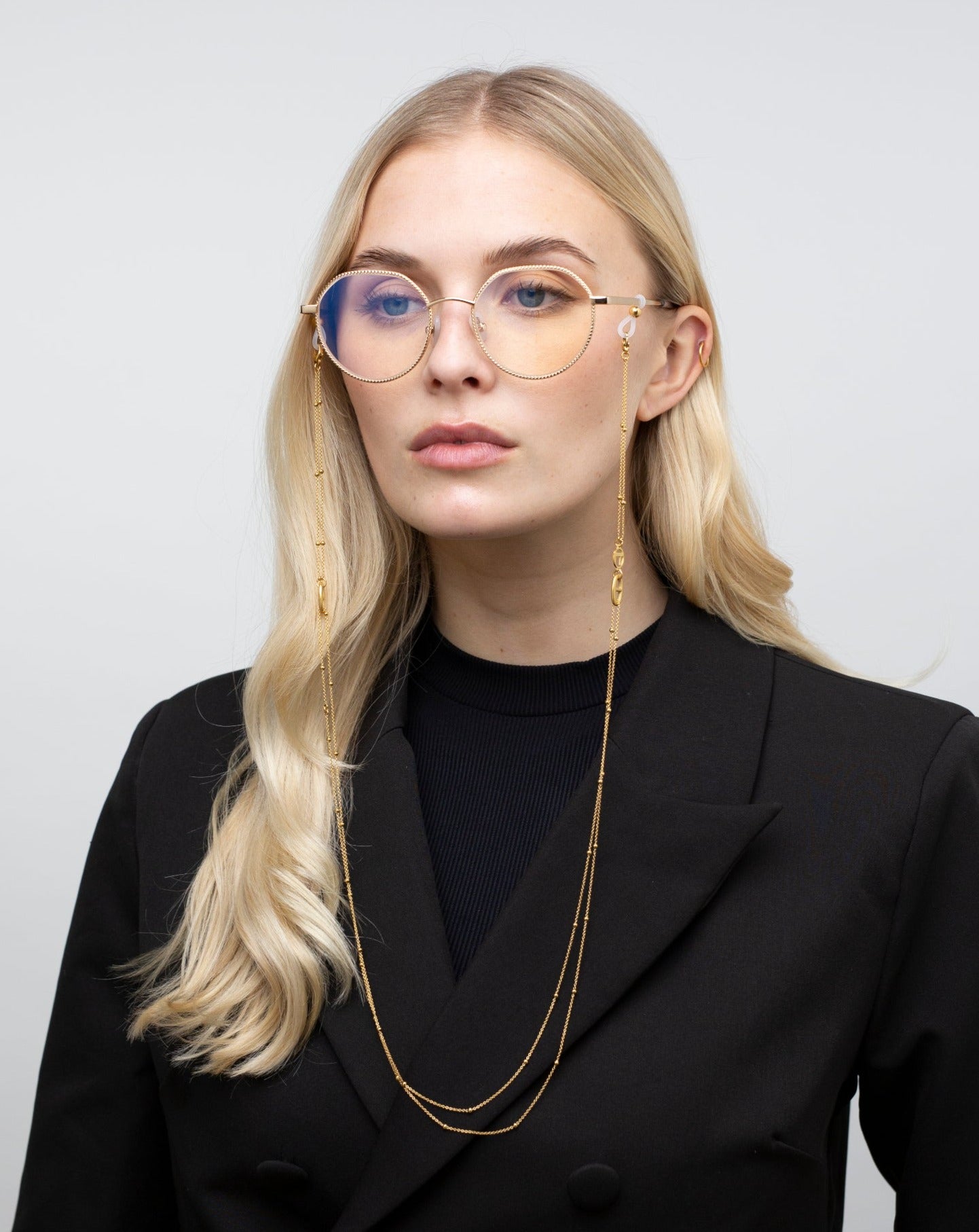 A person with long blonde hair wearing round glasses with a gold chain standing against a plain background. They are dressed in a black blazer over a dark shirt. Their expression is neutral, and they are looking slightly upward.