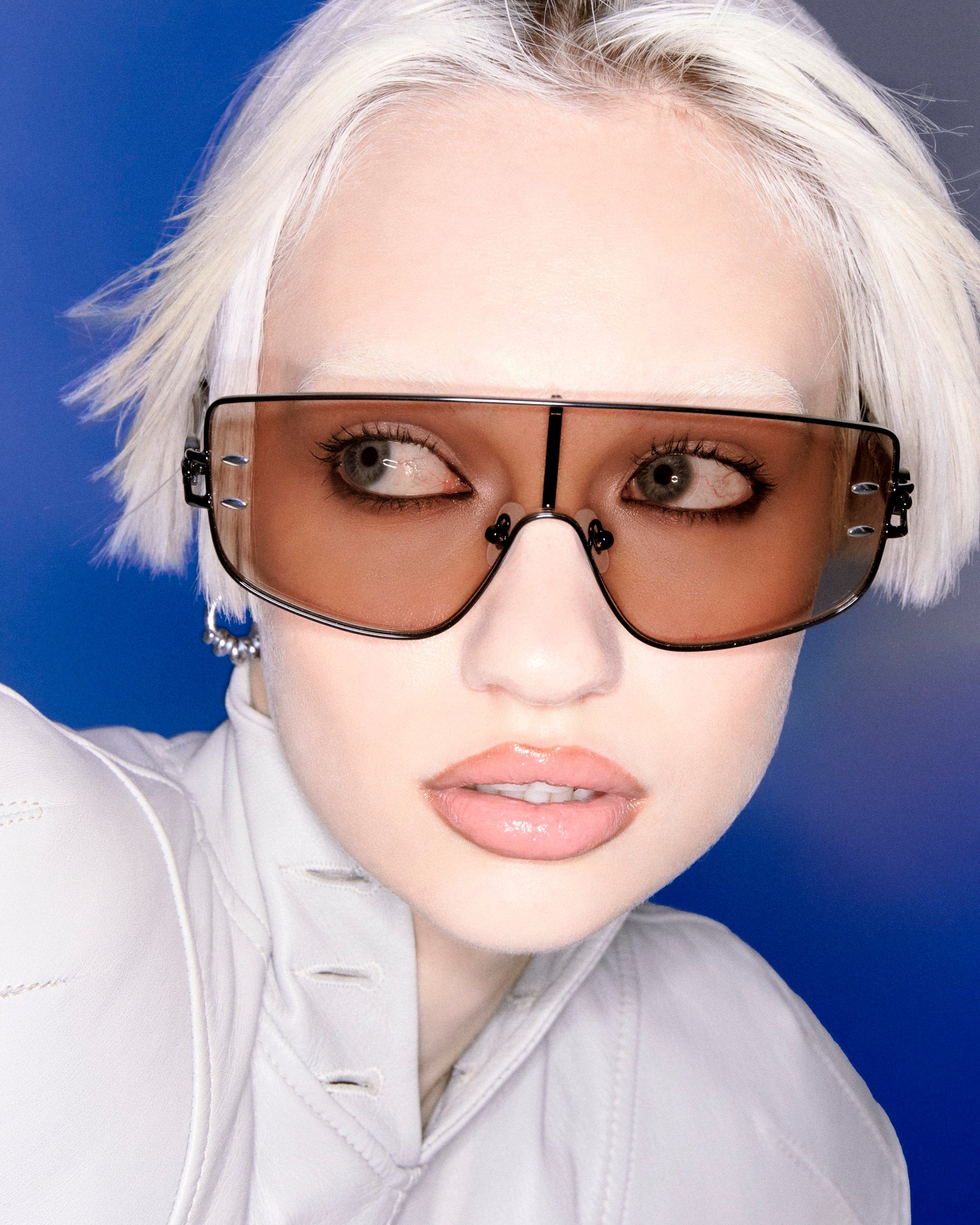 A person with short, platinum blonde hair is wearing large, brown-tinted sunglasses and a white, high-collared top. The background is a gradient of blue and purple hues. The person is gazing to the side with a neutral expression.