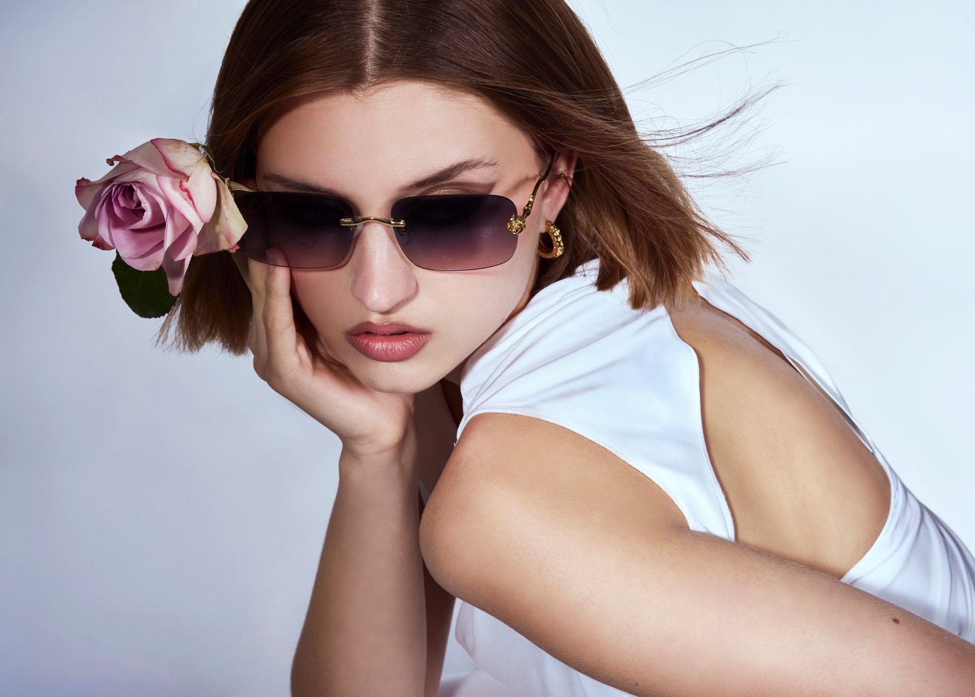 A woman wearing sunglasses and a white dress poses with a pink rose tucked behind her ear, resting her chin on her hand against a plain background.