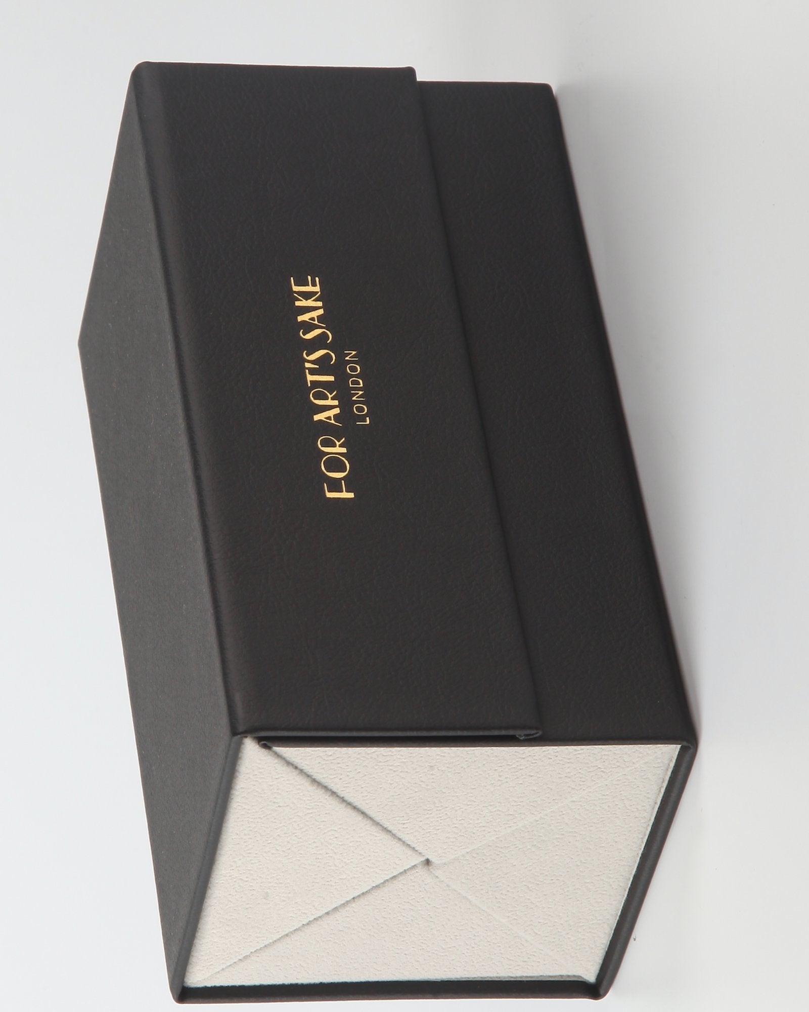 A black, rectangular box with a gold logo on the lid that reads "FOR ART'S SAKE LONDON." The box is open, revealing a white interior with geometric folds. The image is taken from a top angle, showing the exterior and interior simultaneously.