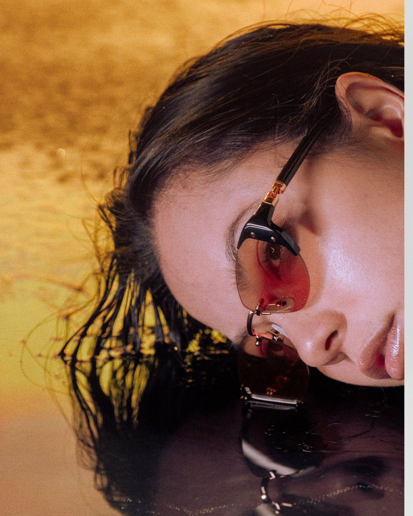 Close-up of a person lying down with their face partially submerged in water. They are wearing black-framed, tinted sunglasses and have long, wet hair. The background is a golden hue, creating a reflective surface around them.