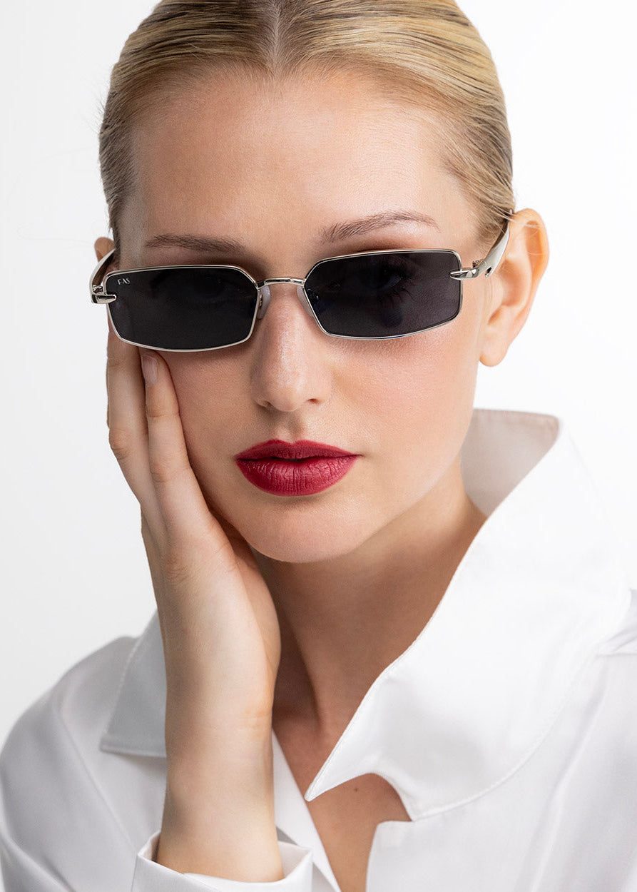Two fashionably dressed individuals wearing glasses. One with black hair and bangs, and the other with platinum blonde hair. Both appear to be posing confidently, showcasing bold makeup and stylish earrings against a plain background.
