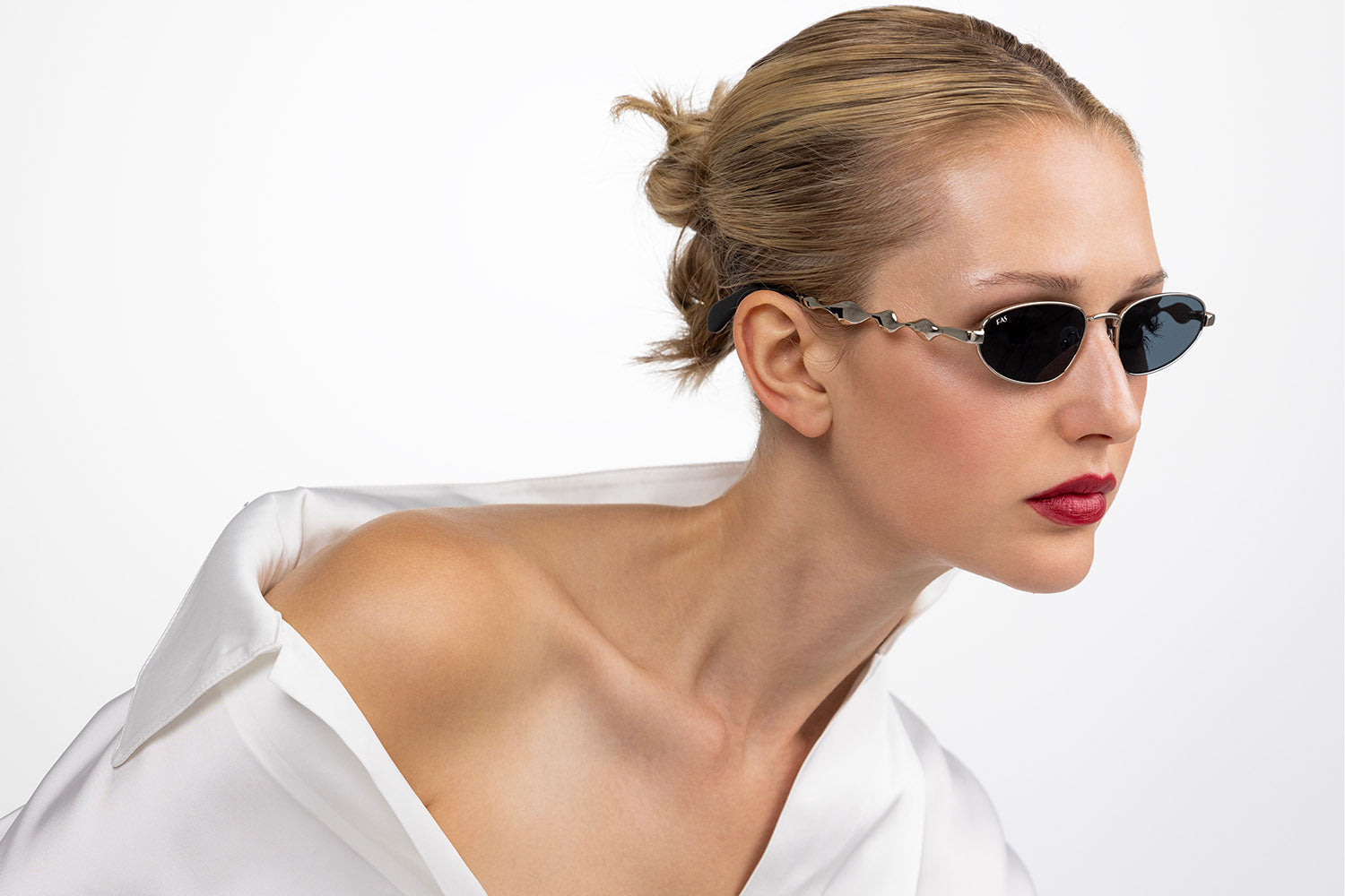 Two women in black outfits wearing sunglasses. The one in the background has a bow and arrow aimed at an apple on the head of the woman in the foreground. The background is white and minimalist.