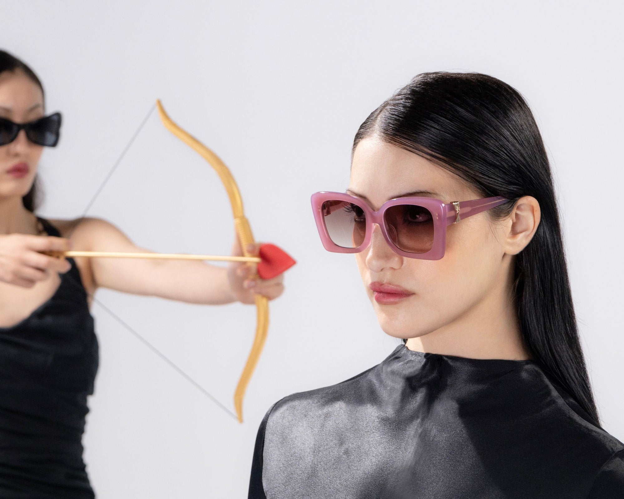 Two women in black outfits wearing sunglasses. The one in the background has a bow and arrow aimed at an apple on the head of the woman in the foreground. The background is white and minimalist.