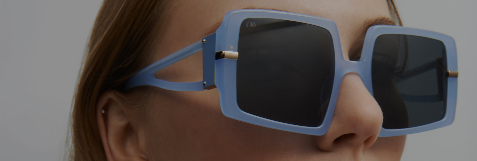 Close-up of a person wearing large, square-shaped sunglasses with pale blue frames and dark lenses. Their brown hair is partially visible, and they have a small ear piercing. The background is a plain, light gray.
