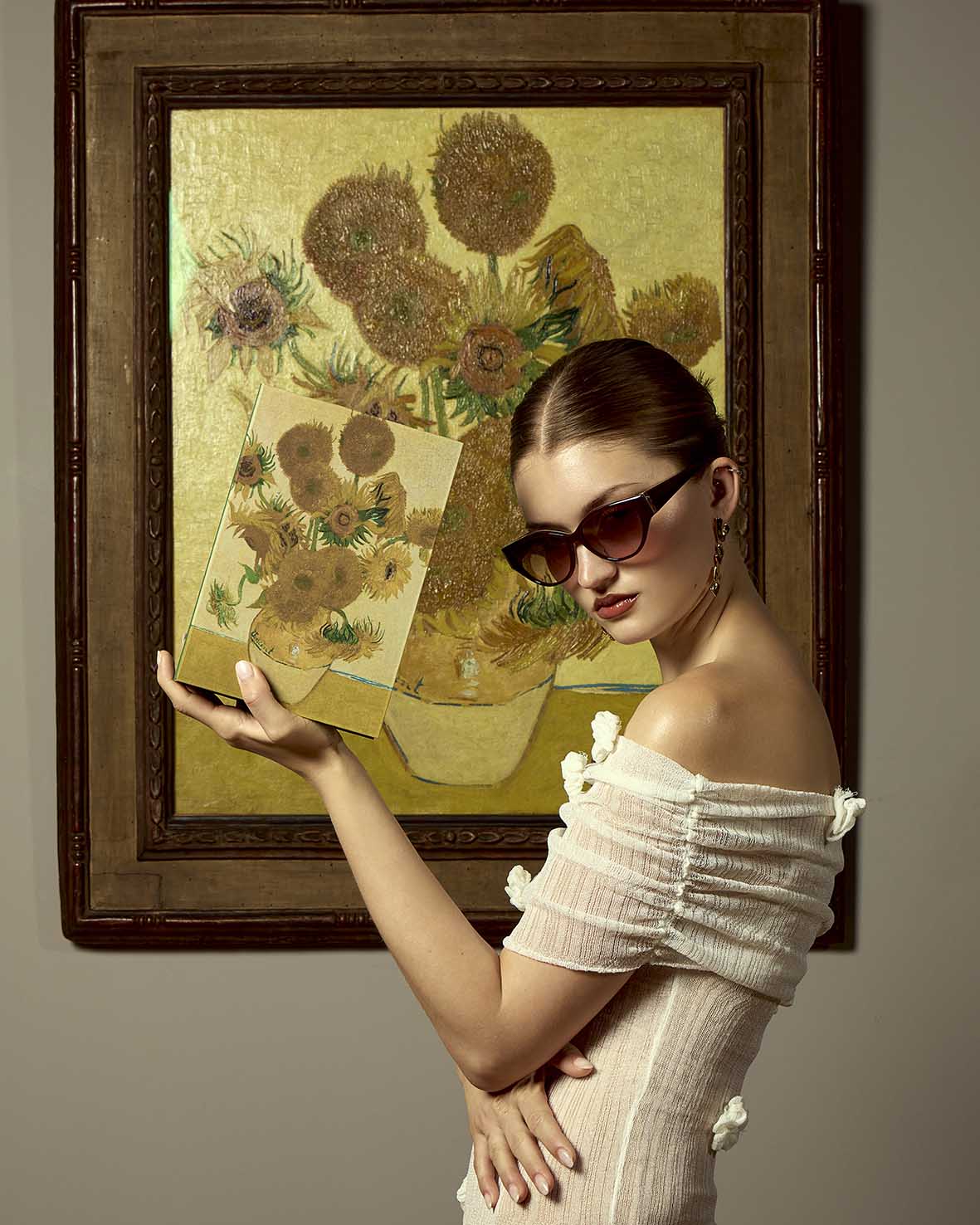 A woman in sunglasses and a white dress poses in front of a sunflower painting, holding a book featuring the same artwork, mirroring the painting behind her.