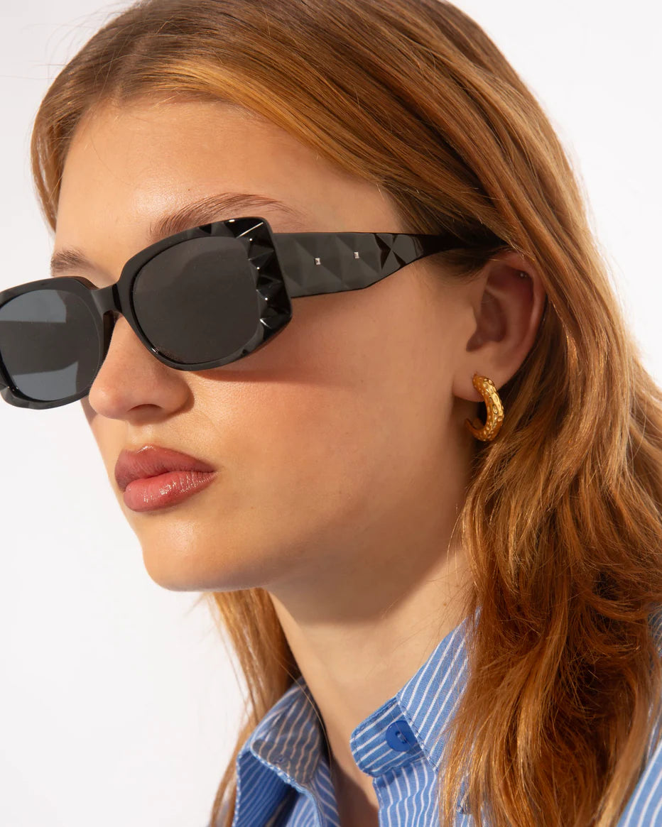A woman with long light brown hair wears black rectangular sunglasses with a geometric pattern on the frame and gold hoop earrings. She is dressed in a blue and white striped shirt and looks off to the side with a neutral expression.