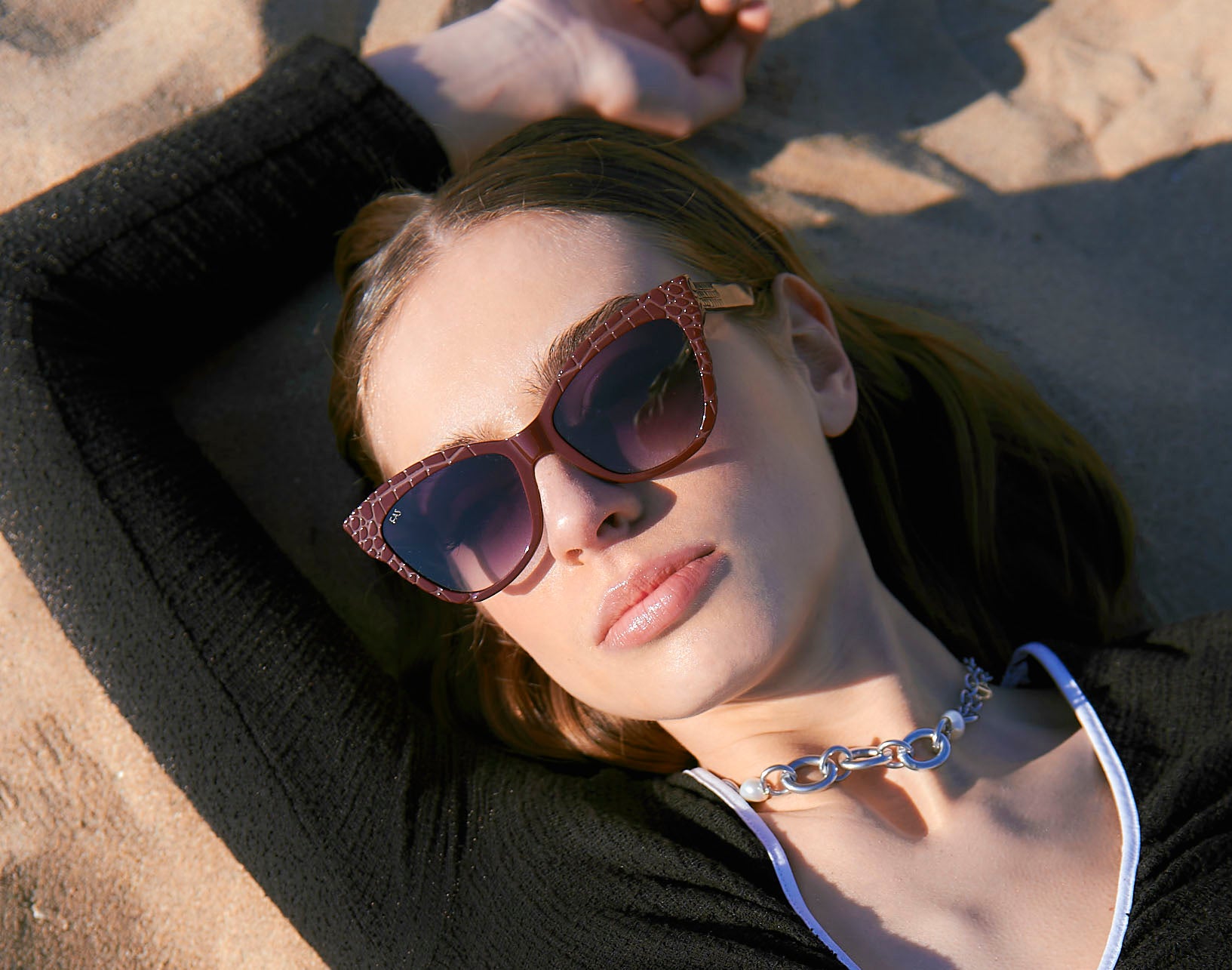 A person wearing stylish sunglasses and a black top with a necklace lies on sandy ground, looking relaxed and serene under the sun.