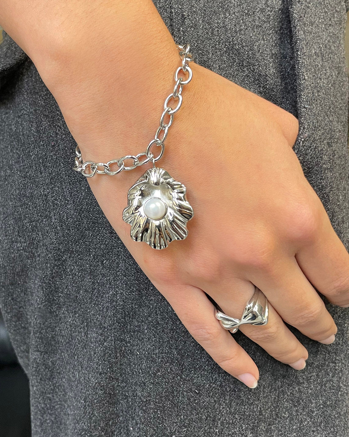 A hand wears The Shell Pendant Bracelet by For Art's Sake®—a chunky silver piece with a freshwater pearl pendant, paired with a matching ring, shown on gray textured fabric from our jewellery collection.