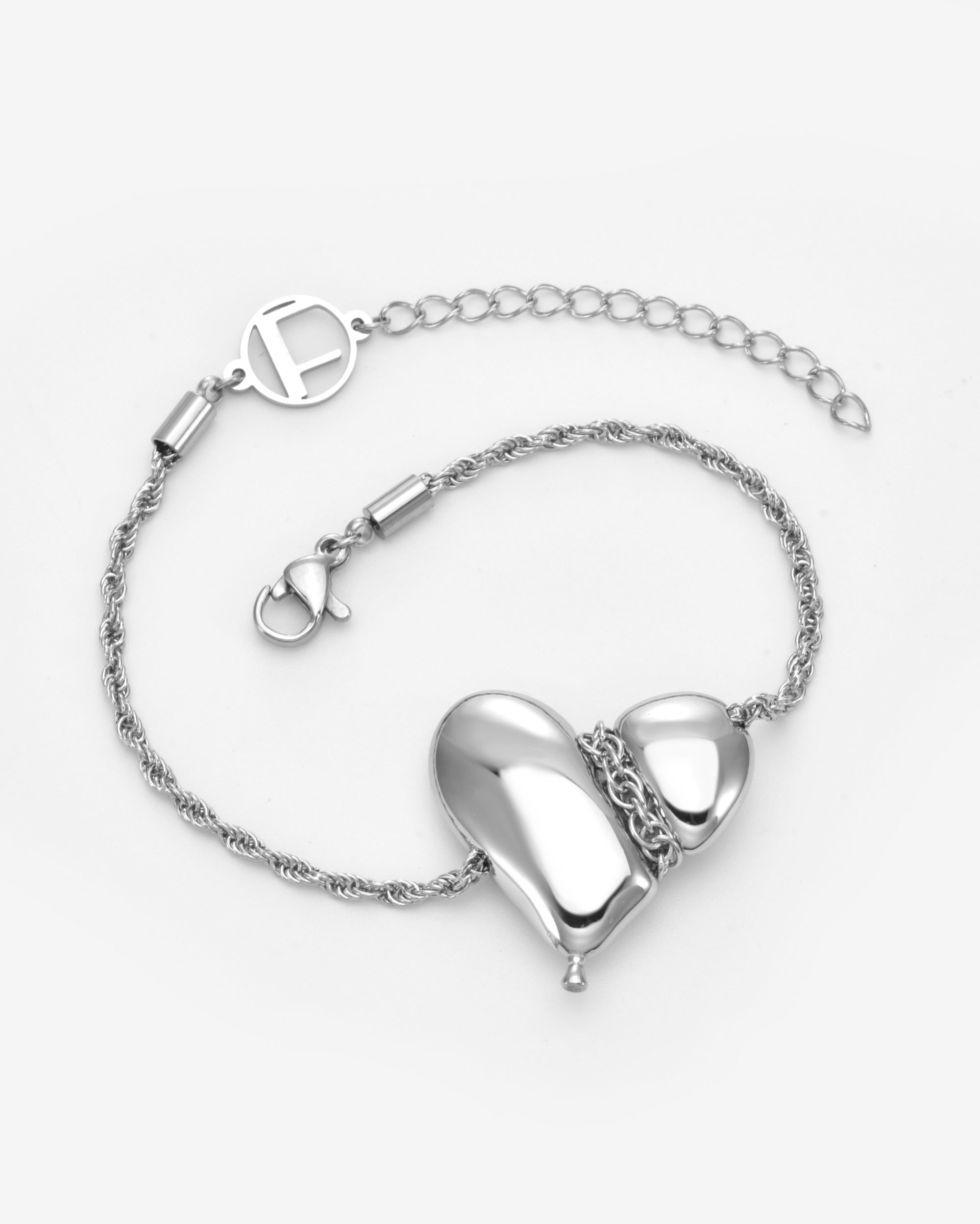 The Puffy Heart Bracelet by For Art's Sake® is a silver chain bracelet with a shiny, abstract puffed heart pendant and an adjustable clasp featuring a circular FAS charm, shown on a white background.