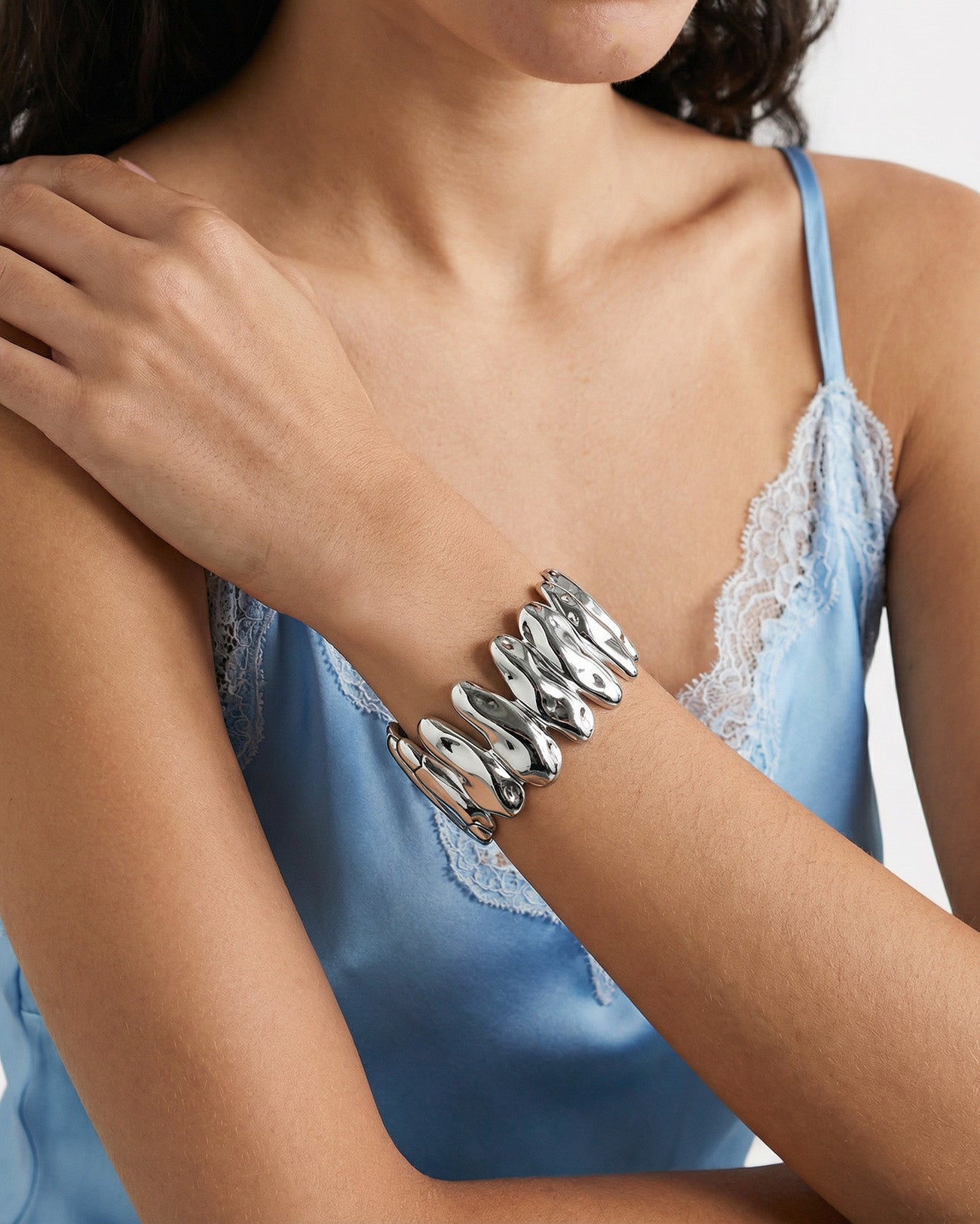 A person wearing a sleeveless light blue satin top with lace trim poses with crossed arms, showcasing The Forms Bangle by For Art's Sake®—a bold, sculptural modern accessory on their wrist.