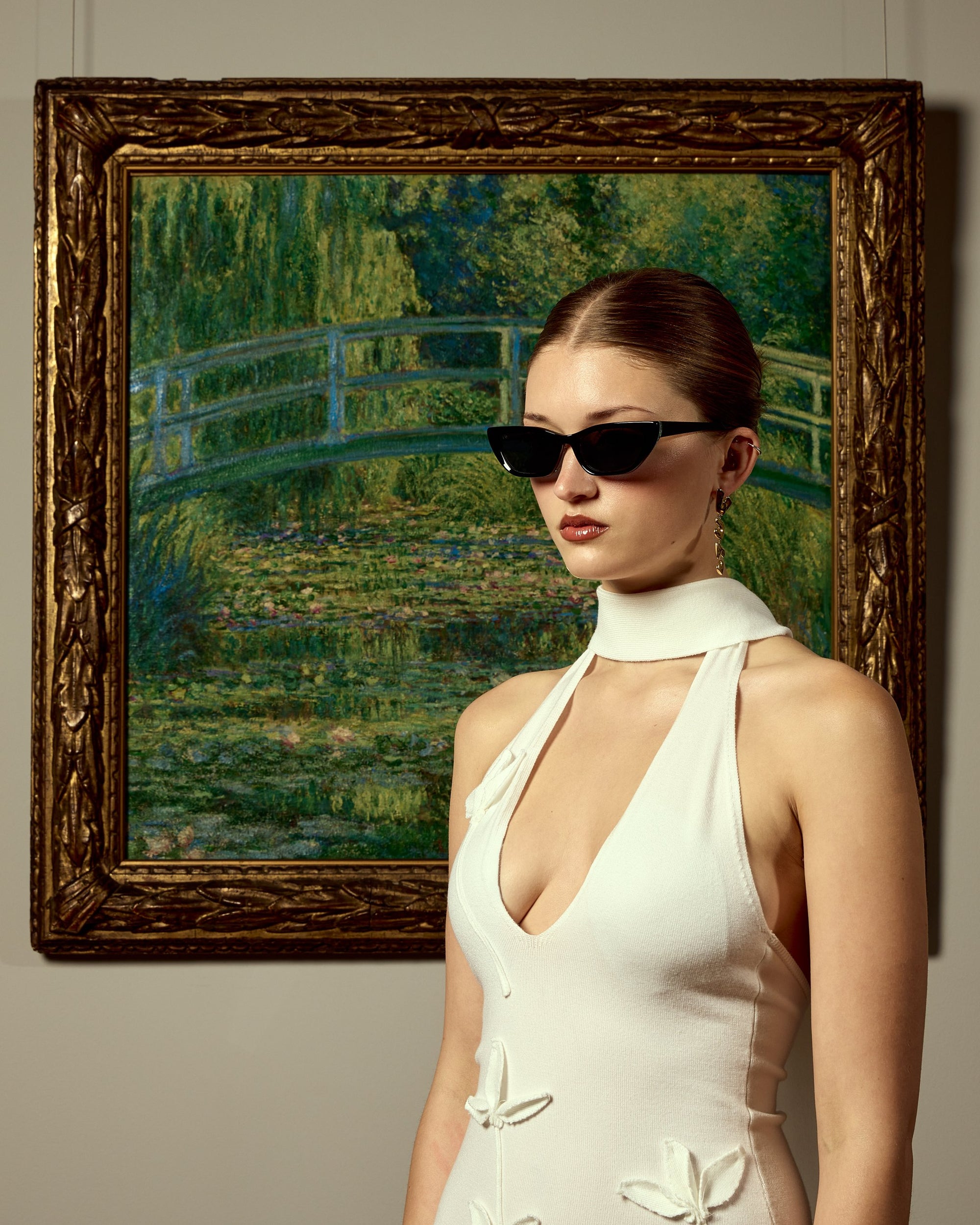 A woman in a white halter dress and For Art's Sake® Claude Monet (Limited Edition) sunglasses stands before a framed painting of a garden bridge over a pond, evoking Monet's Water Lilies.