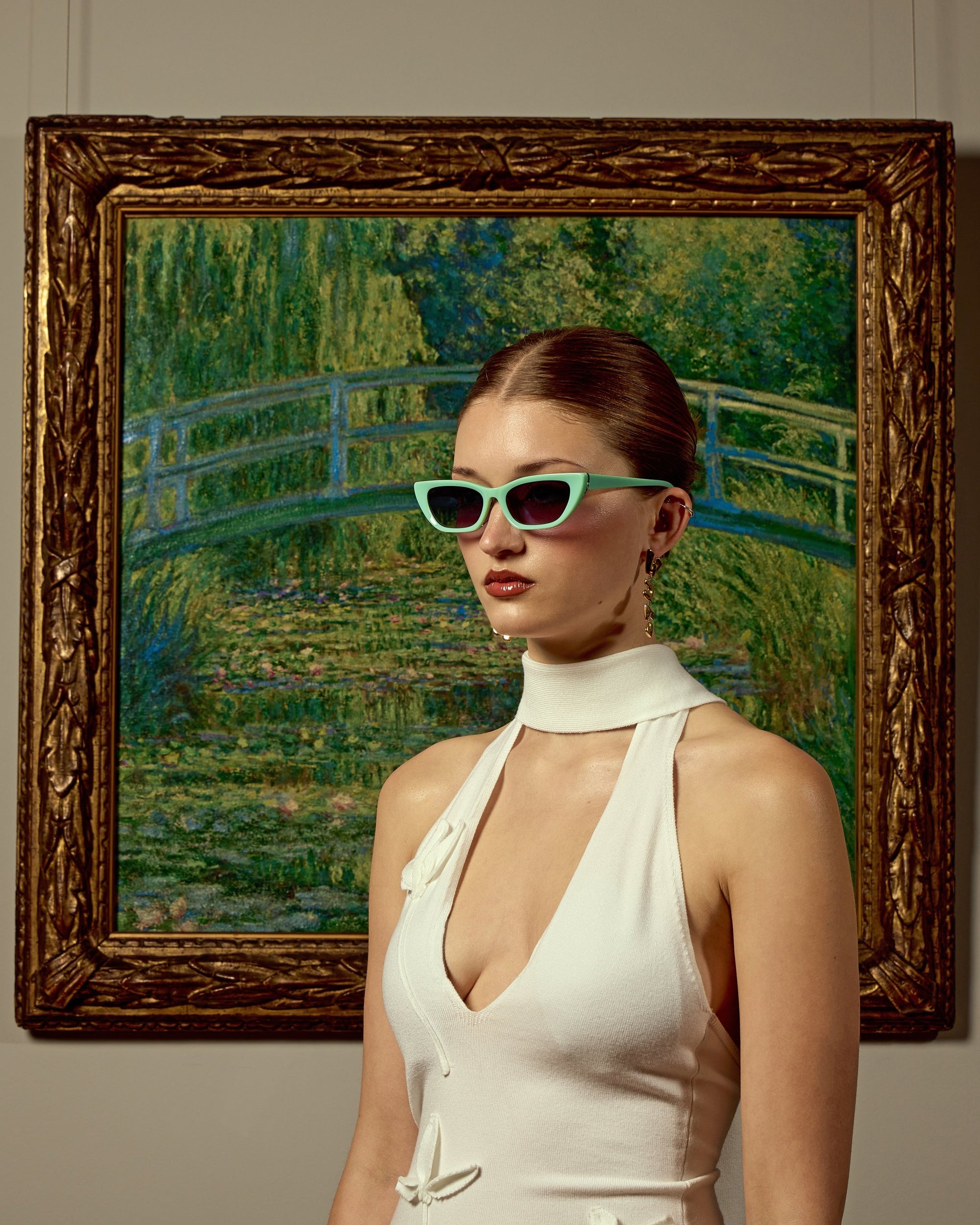 A woman in a chic white dress wears For Art's Sake® Claude Monet (Limited Edition) sunglasses, standing before a framed impressionist painting of a lush garden and arched bridge inspired by Monet.