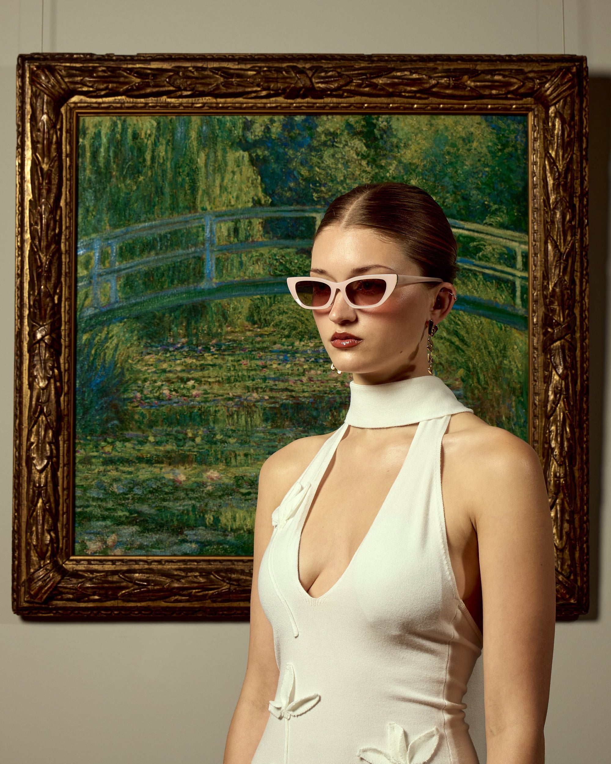 A woman wearing For Art’s Sake® Claude Monet (Limited Edition) sunglasses and a white dress stands before a framed Monet Japanese Bridge, blending modern style with timeless art.