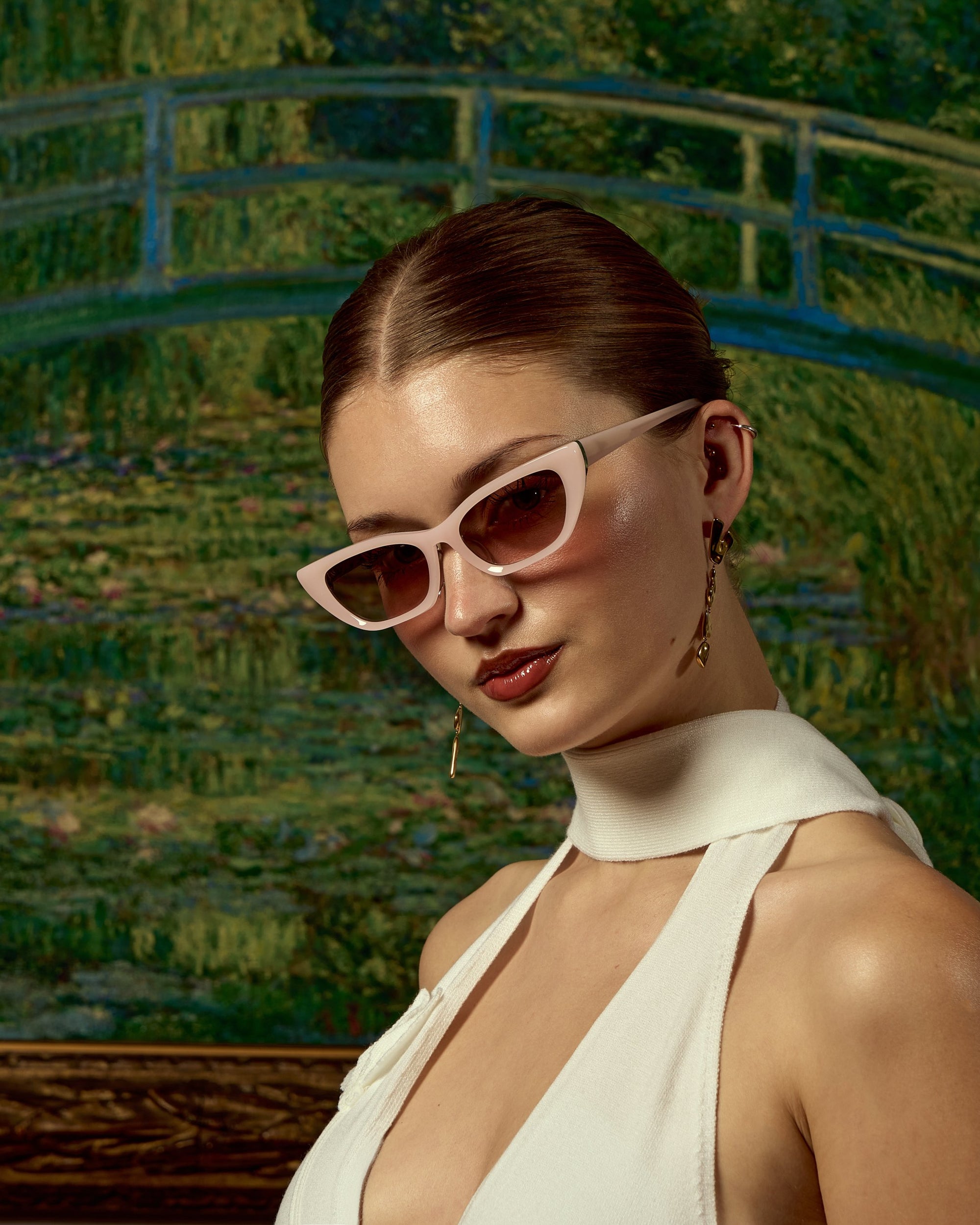 A stylish woman in For Art&#39;s Sake® Claude Monet (Limited Edition) cat-eye sunglasses and a white halter dress poses before Monet’s Japanese Bridge, her hair slicked back and gold earrings glinting.