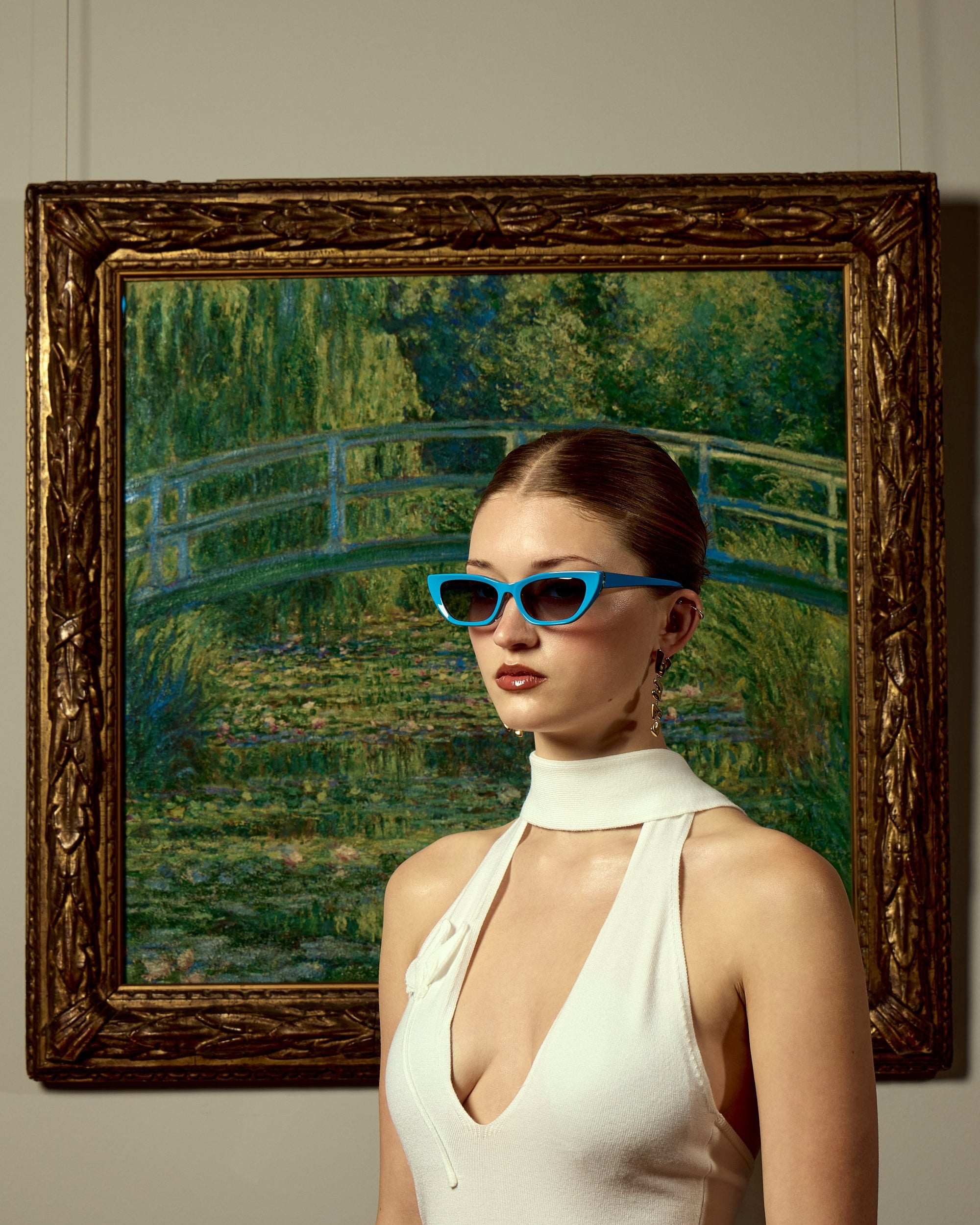 A woman in a white sleeveless dress wears For Art's Sake® Claude Monet (Limited Edition) sunglasses, posing before a framed Monet-style garden bridge painting—fusing modern fashion with classic art.