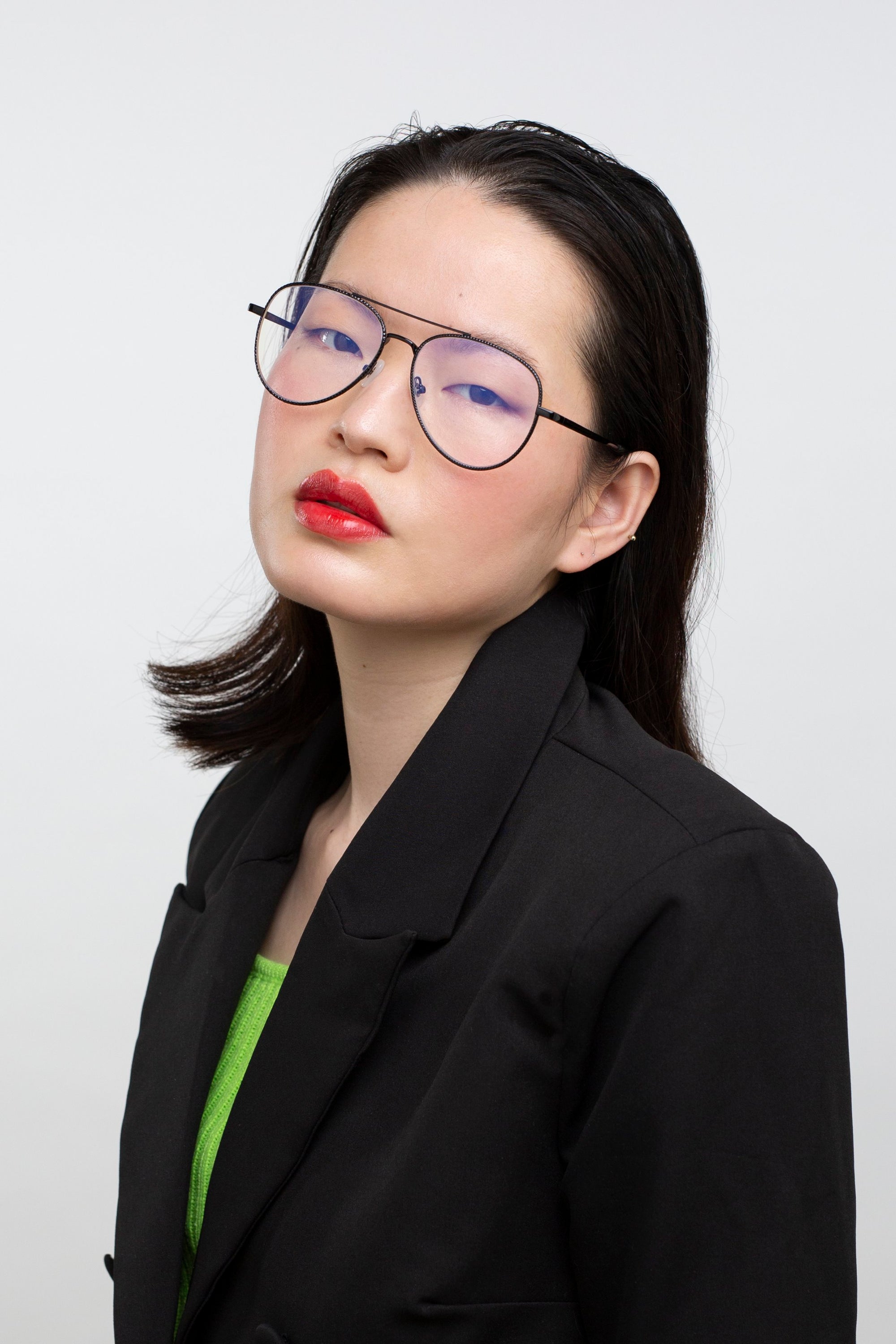 A person with straight dark hair wears the Cleo glasses by For Art's Sake®, a black blazer, and red lipstick, posing confidently against a plain white background. Final sale—no returns accepted.
