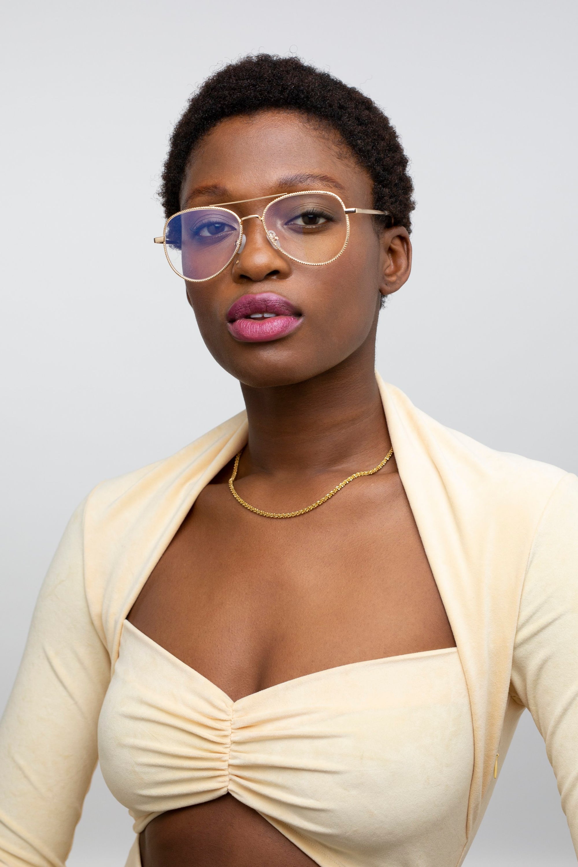 A woman with short curly hair, round glasses, gold necklace, and a light beige Cleo ruched crop top with matching shrug by For Art's Sake® poses against a plain background. Final sale—no returns or reselling allowed.
