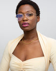 A woman with short curly hair, round glasses, gold necklace, and a light beige Cleo ruched crop top with matching shrug by For Art's Sake® poses against a plain background. Final sale—no returns or reselling allowed.