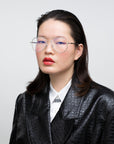 A person with shoulder-length dark hair, wearing the Cleo round, wire-framed optical glasses from For Art's Sake® and red lipstick, is dressed in a black textured jacket over a white shirt. The individual has a neutral expression and stands against a plain white background.