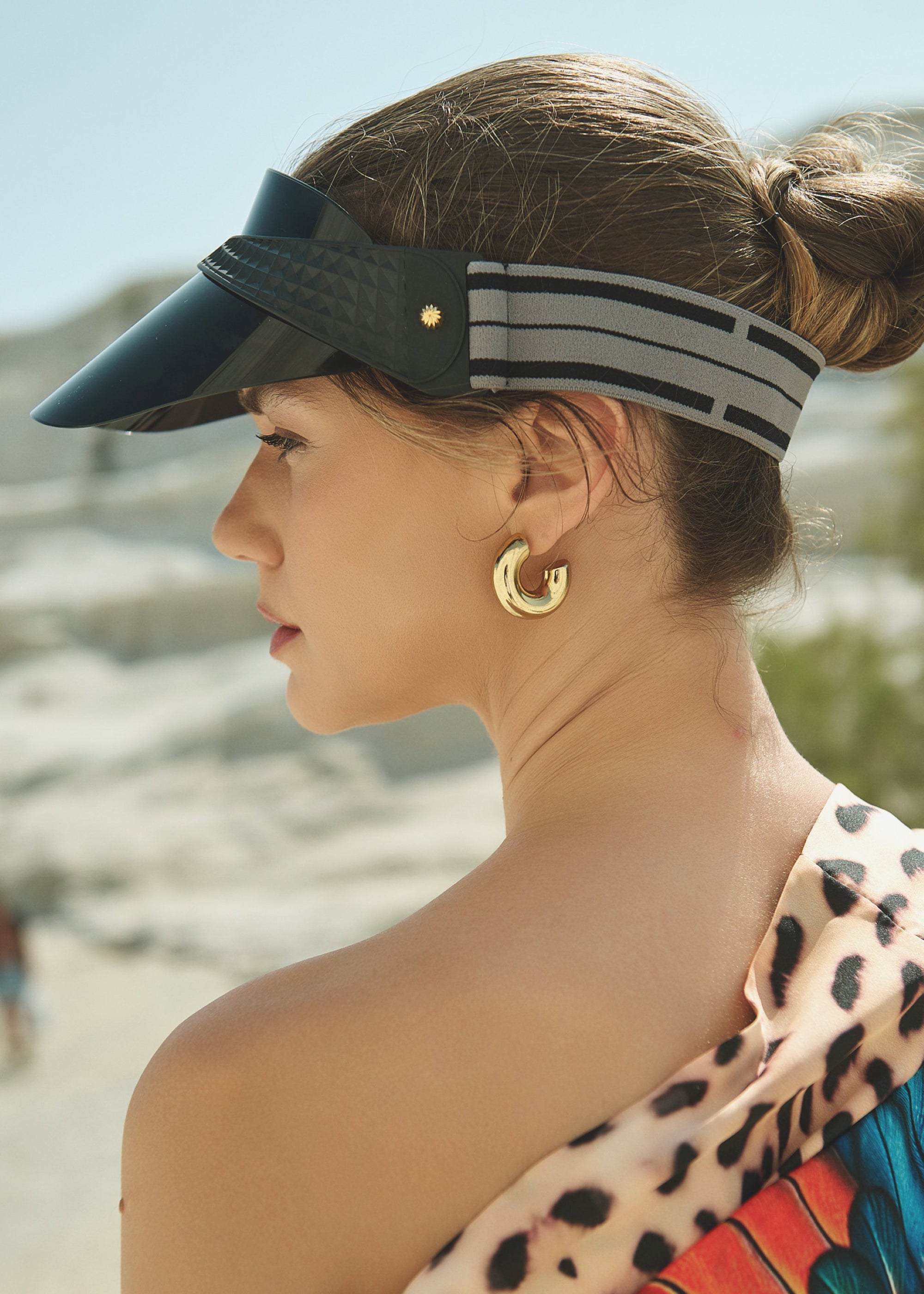 A woman wears the For Art's Sake® Cloud Visor (UV protected, black) and gold hoop earrings with her hair in a low bun. She dons a colorful leopard print garment and stands outdoors, looking off to the side in bright sunlight.