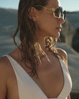 A woman with wet hair wears For Art's Sake® Cry Me a River sunglasses, featuring gold details, paired with a white swimsuit and posing in sunlit outdoors against blurred rocks.