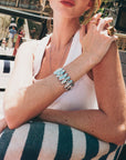 A woman in a white tank top and striped cushion showcases For Art's Sake® The Forms Bangle on her wrist, sunlight highlighting her freckled skin.