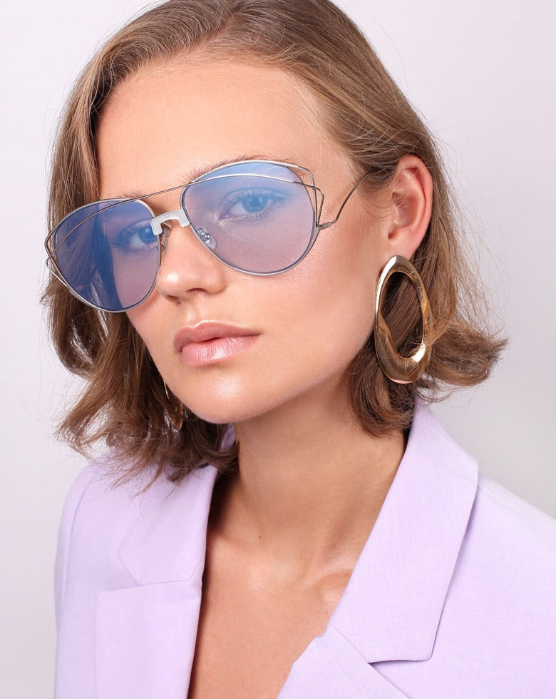 A person with short, wavy hair is wearing the Dark Eyes sunglasses by For Art's Sake®, featuring large, thin frames with blue-tinted nylon lenses. They complement their look with oversized gold hoop earrings and a light purple blazer while posing against a plain, light background.