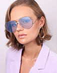 A person with short, wavy hair is wearing the Dark Eyes sunglasses by For Art's Sake®, featuring large, thin frames with blue-tinted nylon lenses. They complement their look with oversized gold hoop earrings and a light purple blazer while posing against a plain, light background.
