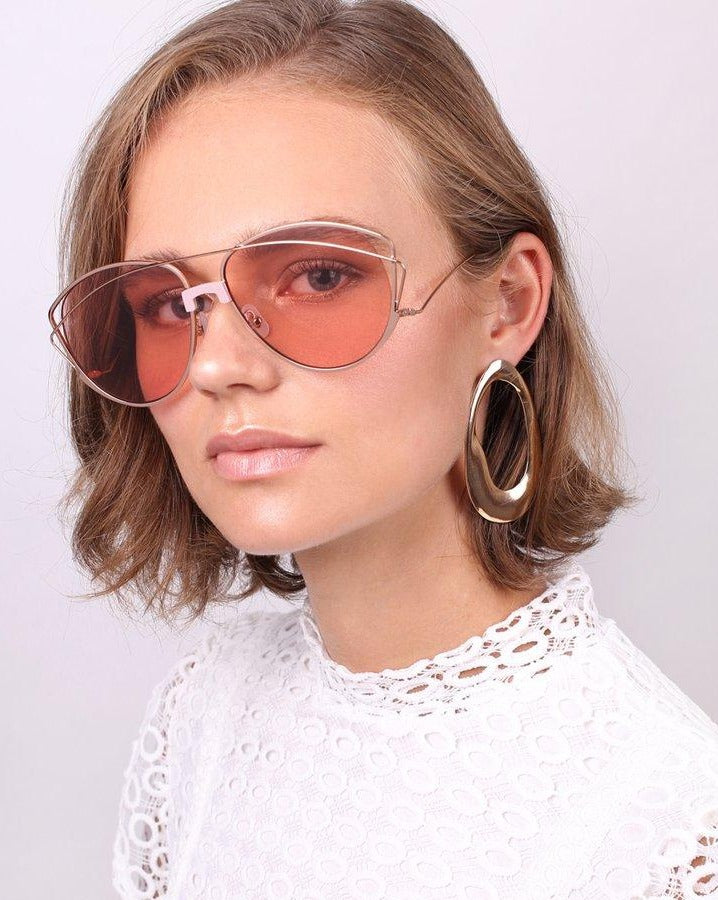 A person with short, light brown hair is wearing the Dark Eyes large pink-tinted sunglasses with stainless steel frames from For Art's Sake®, along with oversized hoop earrings. They are dressed in a white lace top and are looking directly at the camera with a neutral expression against a plain, light-colored background.