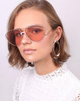A person with short, light brown hair is wearing the Dark Eyes large pink-tinted sunglasses with stainless steel frames from For Art's Sake®, along with oversized hoop earrings. They are dressed in a white lace top and are looking directly at the camera with a neutral expression against a plain, light-colored background.