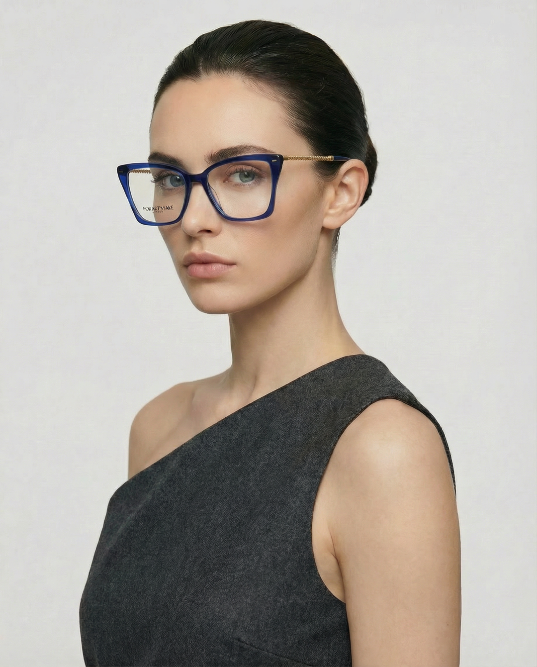A woman with dark hair tied back wears For Art's Sake® Dion large cat-eye glasses and a sleeveless, dark gray one-shoulder dress. She stands slightly turned against a plain light background.
