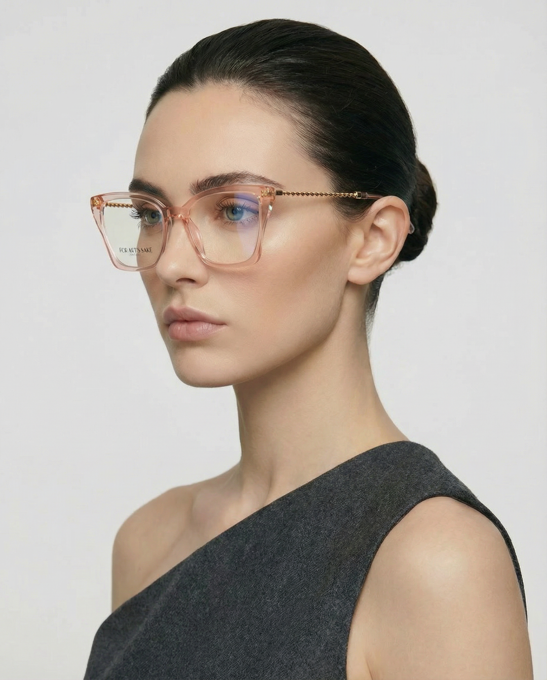 A woman with fair skin and dark hair in a low bun wears For Art's Sake® Dion oversized translucent pink cat-eye glasses with UV protection and a sleeveless asymmetrical gray top, posing against a plain light background.
