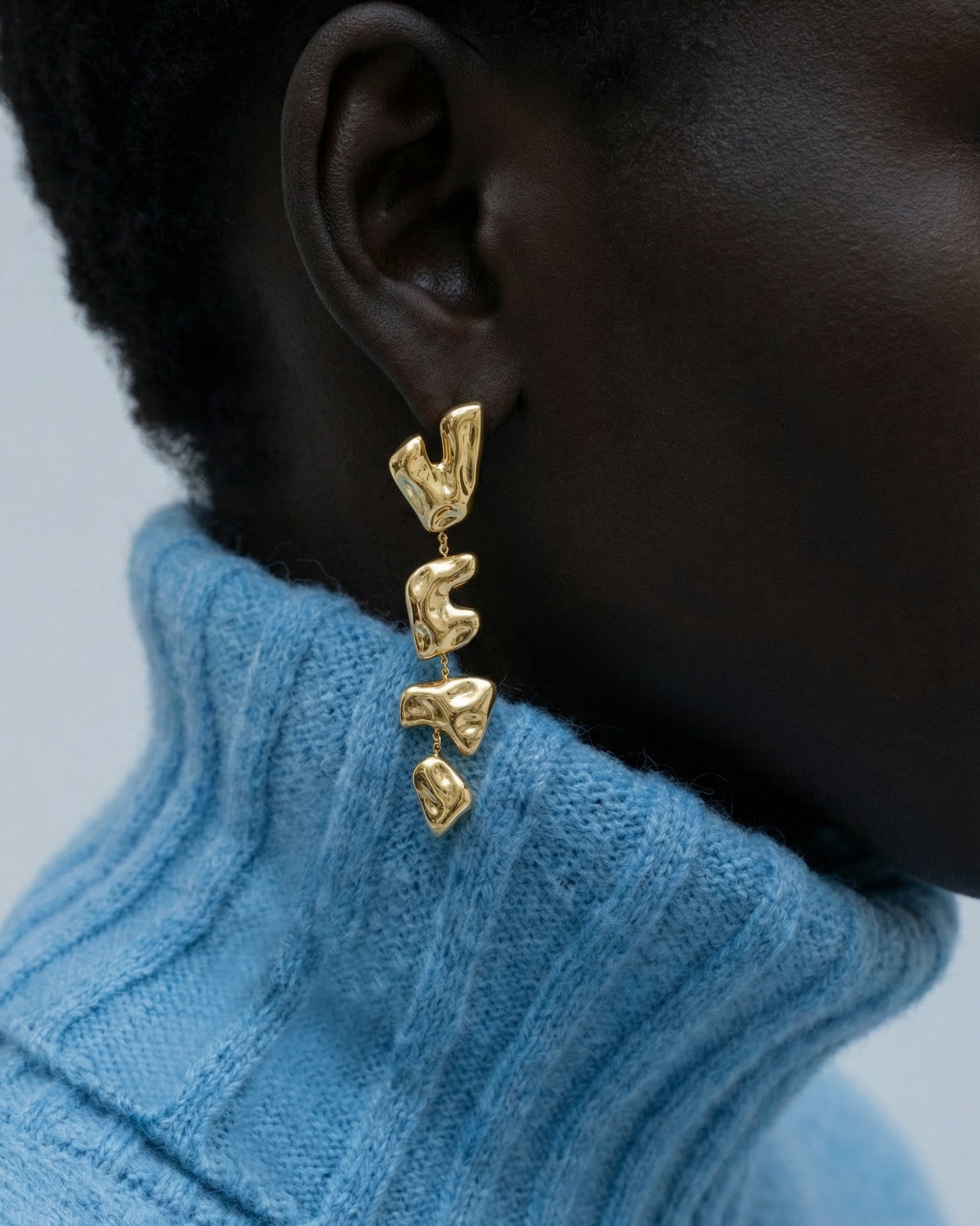 Close-up of a person wearing The Forms Earrings by For Art's Sake®—textured gold abstract dangles—paired with a light blue ribbed turtleneck. The modern jewellery pops against the soft, neutral background.