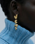Close-up of a person wearing The Forms Earrings by For Art's Sake®—textured gold abstract dangles—paired with a light blue ribbed turtleneck. The modern jewellery pops against the soft, neutral background.