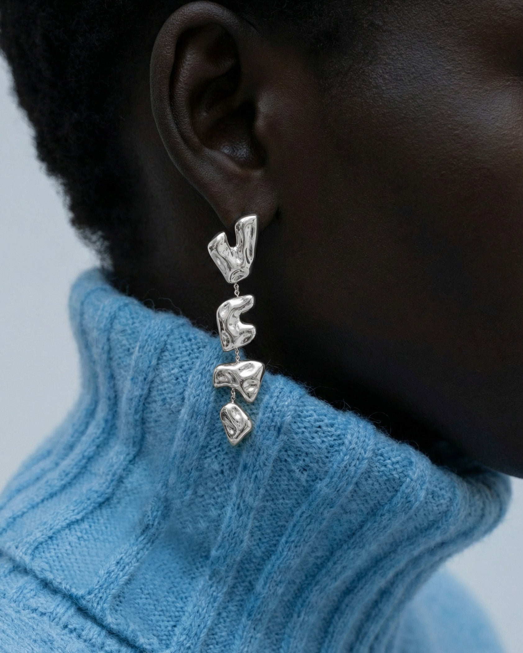 Close-up of a person wearing The Forms Earrings by For Art's Sake®—chunky, abstract silver metal with three segments—paired with a light blue ribbed turtleneck. A softly blurred background highlights the modern style.