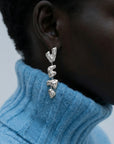 Close-up of a person wearing The Forms Earrings by For Art's Sake®—chunky, abstract silver metal with three segments—paired with a light blue ribbed turtleneck. A softly blurred background highlights the modern style.