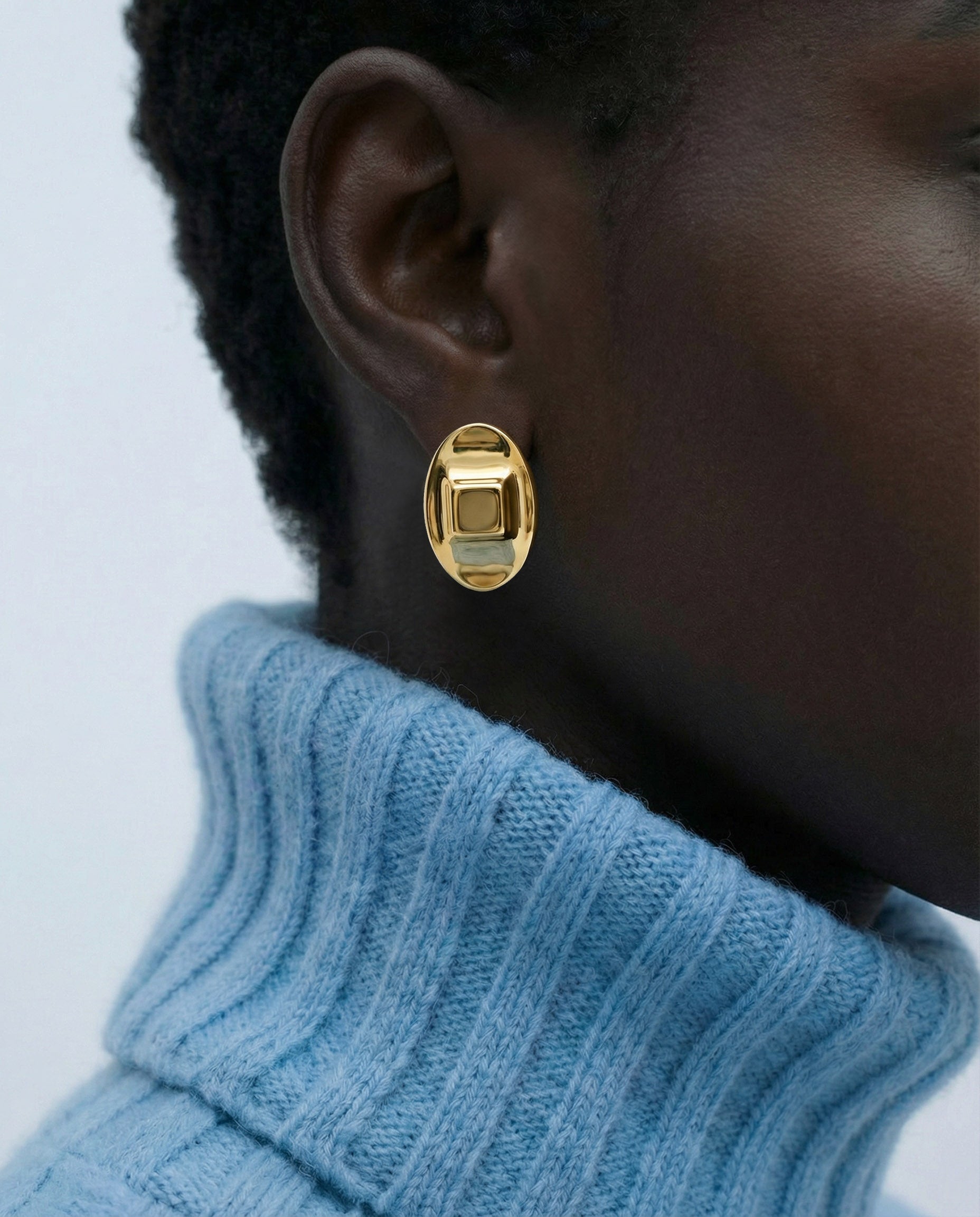 A close-up shows a person wearing The Geometry Earrings by For Art's Sake®—modern, chunky gold earrings—paired with a light blue ribbed turtleneck against a plain background.