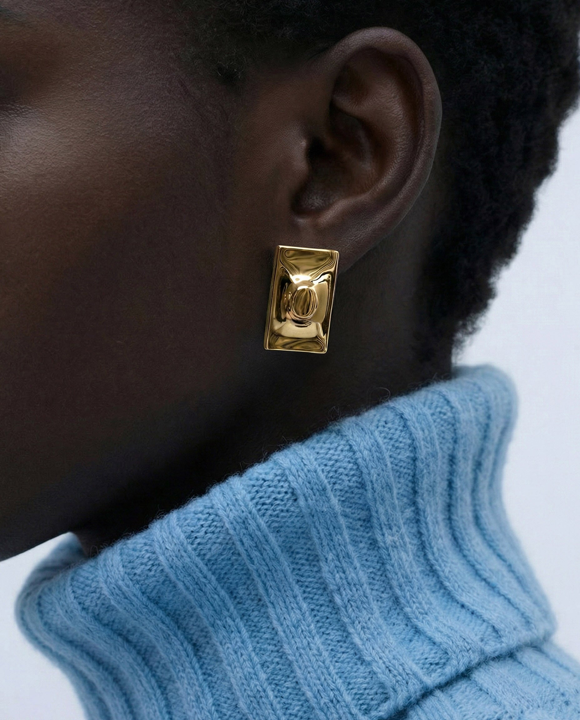 Close-up of a person wearing For Art's Sake® The Geometry Earrings—chunky, rectangular statement earrings—paired with a light blue ribbed turtleneck; only the lower face, ear, and neck are visible to highlight the modern design.