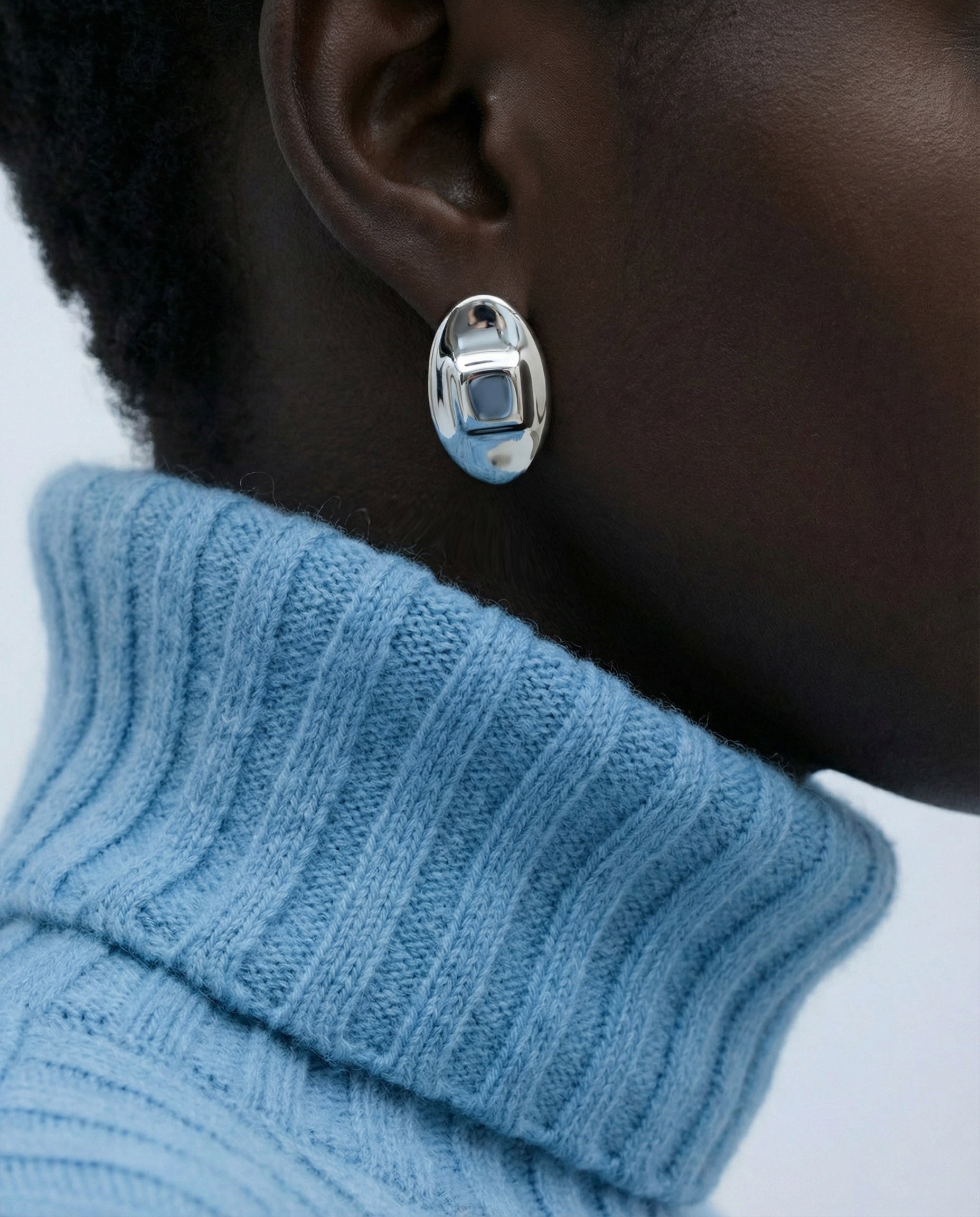 Close-up of a person in a light blue ribbed turtleneck sweater wearing For Art's Sake® The Geometry Earrings—sleek, shiny ovals that stand out against dark skin and a neutral background.