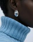 Close-up of a person in a light blue ribbed turtleneck sweater wearing For Art's Sake® The Geometry Earrings—sleek, shiny ovals that stand out against dark skin and a neutral background.
