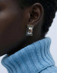 A close-up of a person wearing For Art’s Sake® The Geometry Earrings and a thick, ribbed light blue turtleneck. The plain background highlights the earrings’ modern design and the sweater’s textured fabric.
