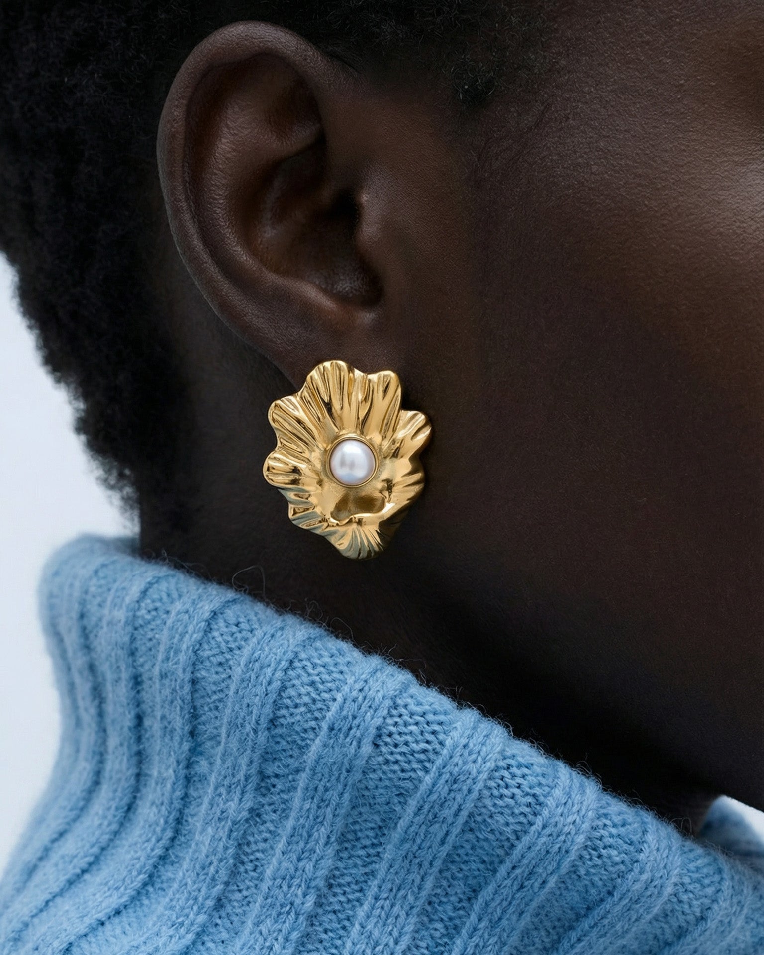 A close-up of a person wearing For Art's Sake® The Shell Earrings—large gold flower-shaped earrings with a freshwater pearl center—paired with a light blue ribbed turtleneck, showcasing an elegant coastal jewelry style.